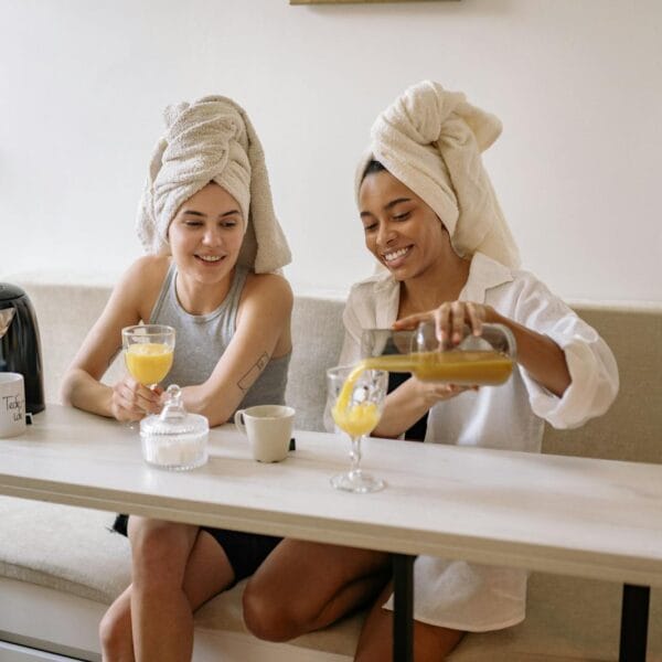 Two friends enjoying a relaxing morning indoors, sharing fresh juice with towels on their heads.